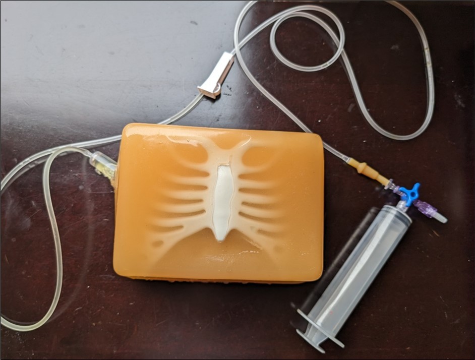 Showing the complete model of the neonatal pericardial effusion. We can see the spiked portion of the sailing bag coming out from the model. We can also see some yellow-colored fluid coming out in the tubing which represents the simulated pericardial effusion fluid. The red arrow shows a three-way stopcock that connects the tubing to a 50 ML syringe. This syringe will be used to refill the saline bag simulating the pericardial effusion once the fluid is decreasing after it's drained by the trainee